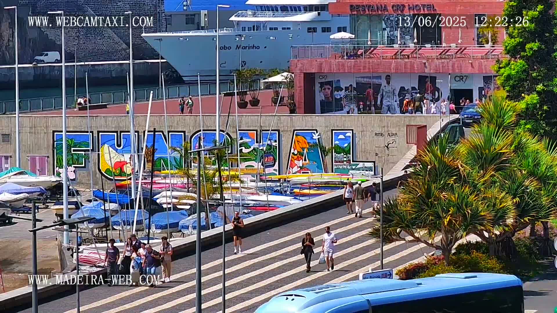 A sunny waterfront scene shows a harbor filled with sailboats and kayaks, a large mural depicting the word "FUNCHAL," pedestrians walking along a striped walkway, and a large cruise ship docked in the background.