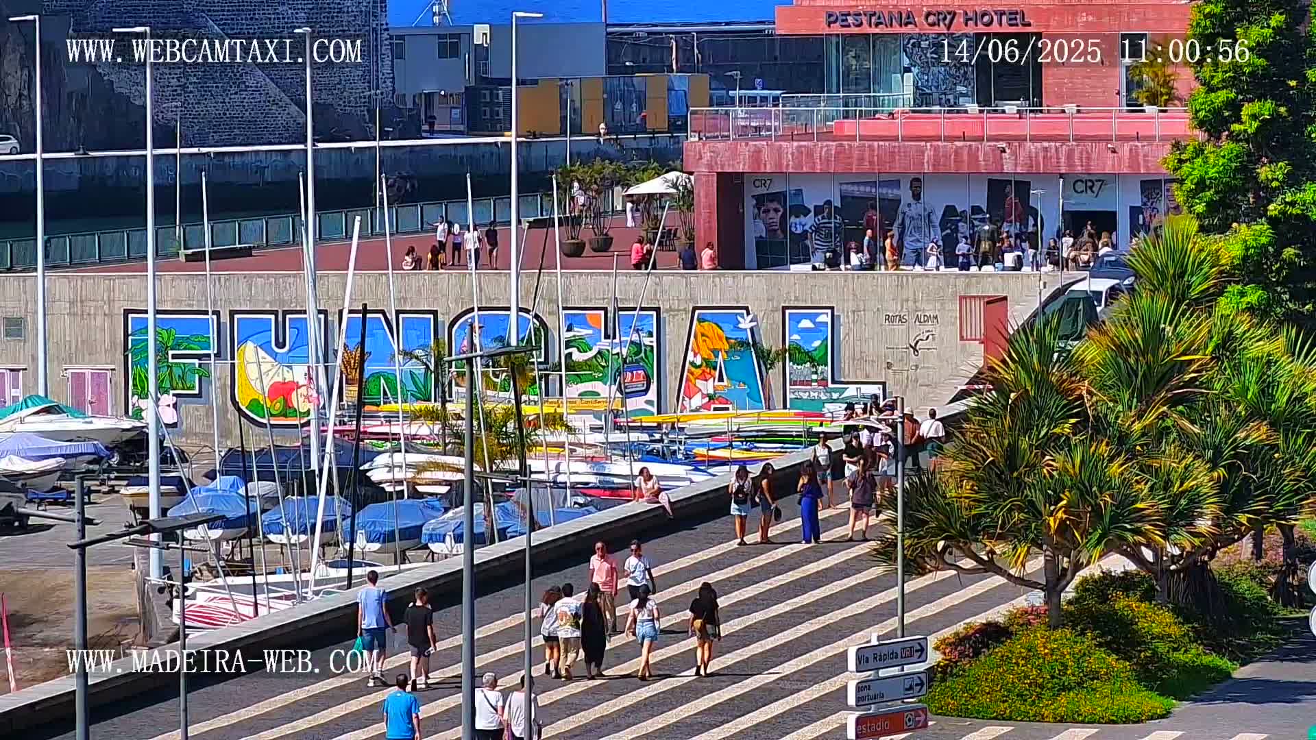 A sunny harbor scene shows a large mural of "FUNCHAL" on a building overlooking a marina filled with sailboats and small motorboats, with numerous people walking along a paved walkway.