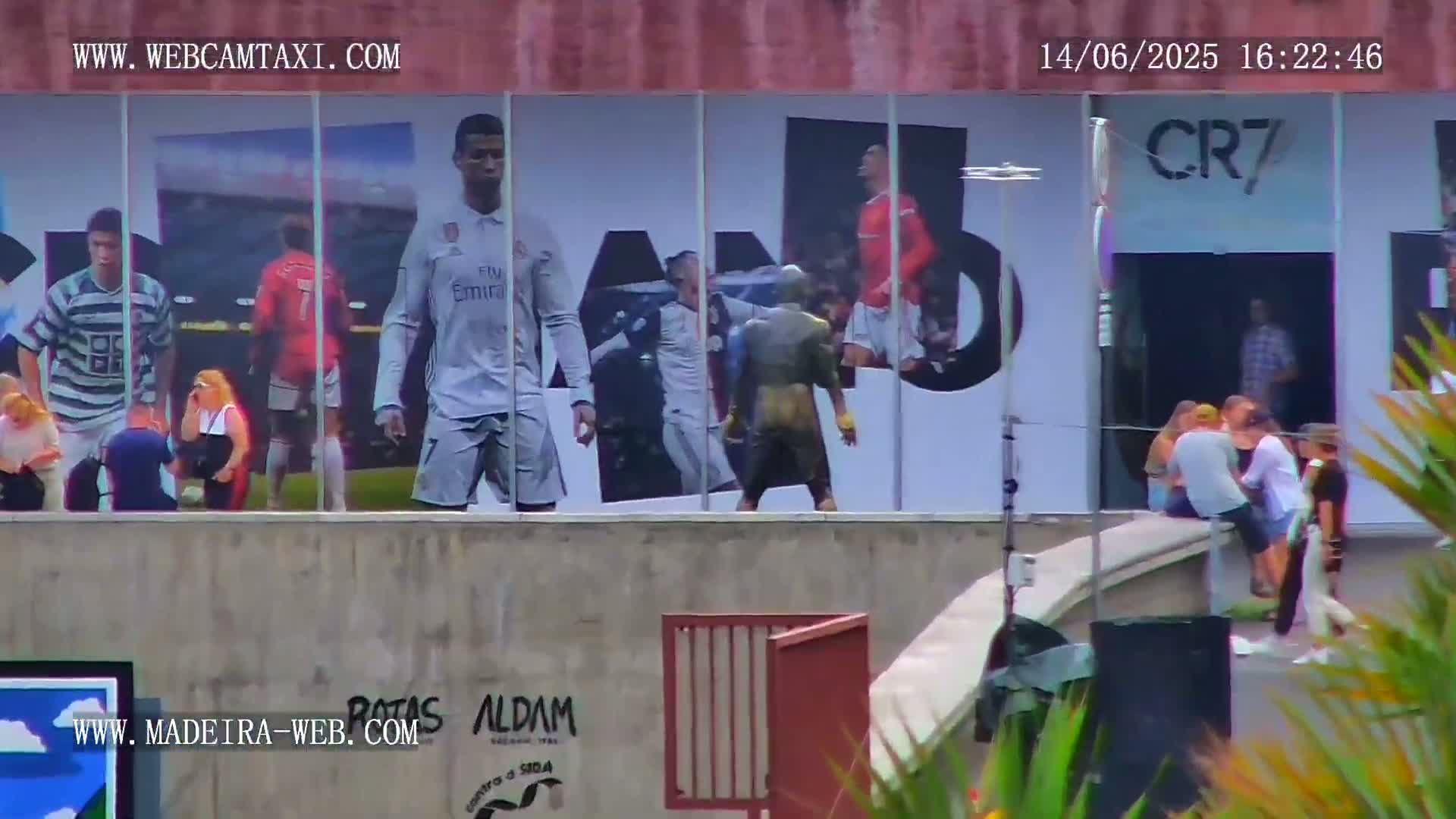 A large building with large images of soccer players on its exterior walls, is surrounded by people, under a partly cloudy sky.