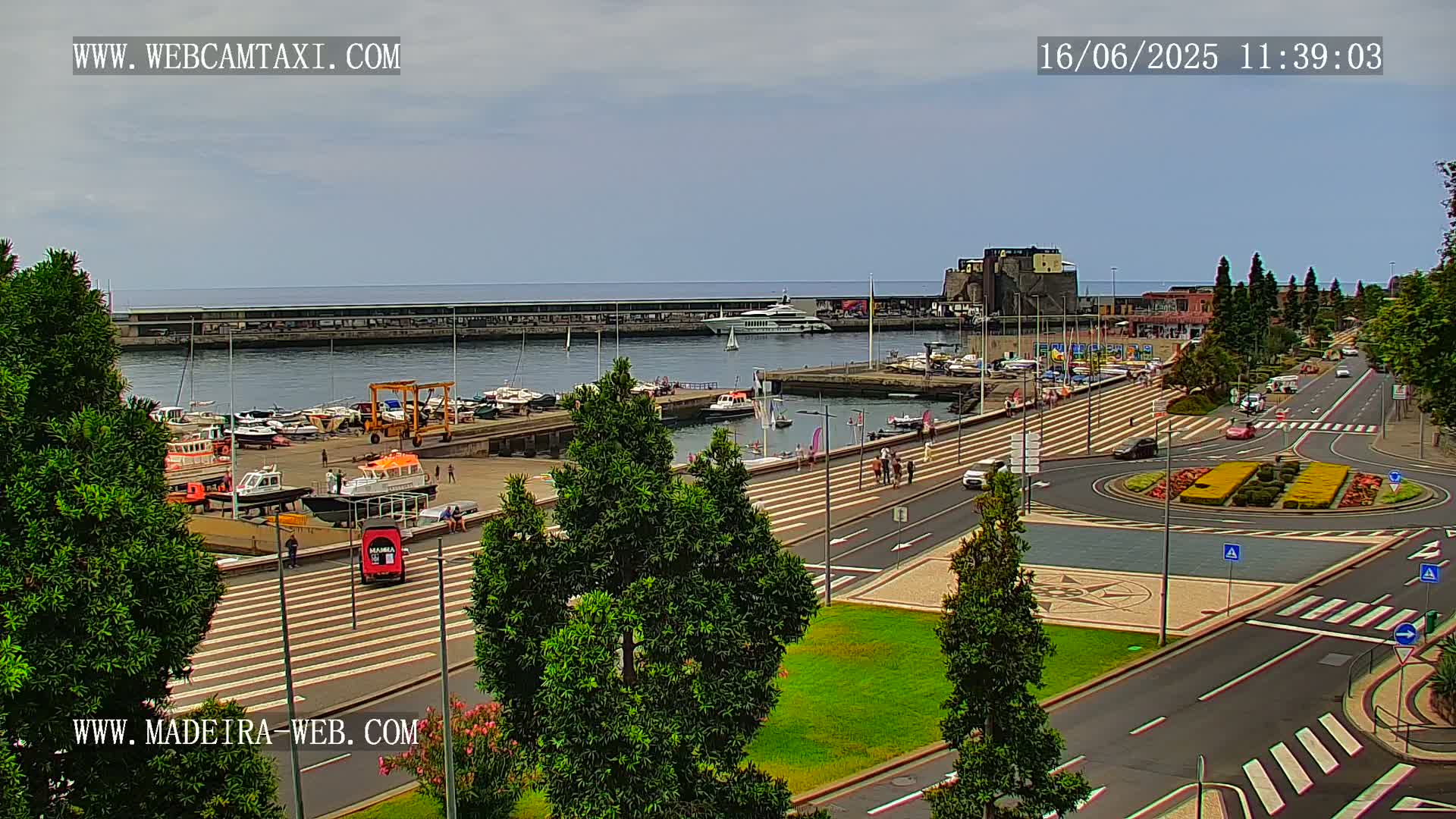 A harbor filled with boats is adjacent to a paved roadway and roundabout with landscaping under a partly cloudy sky.