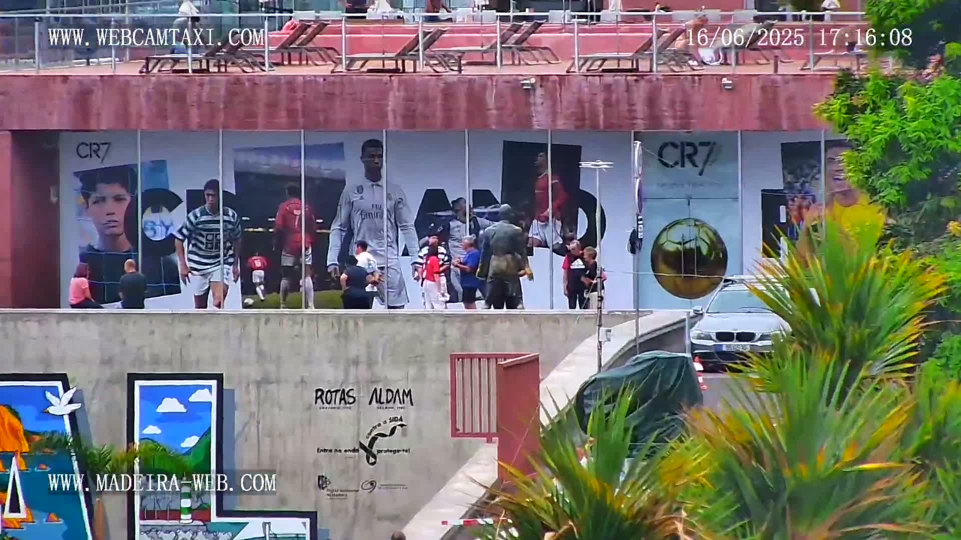 A building displays large images of Cristiano Ronaldo in various soccer jerseys, with people standing in front and a car visible in the lower right corner, partially obscured by lush green plants.