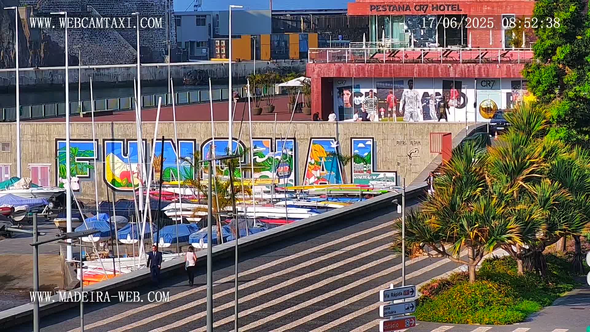 A sunny harbor scene shows numerous boats docked alongside a wall featuring large, colorful murals, with people walking nearby.