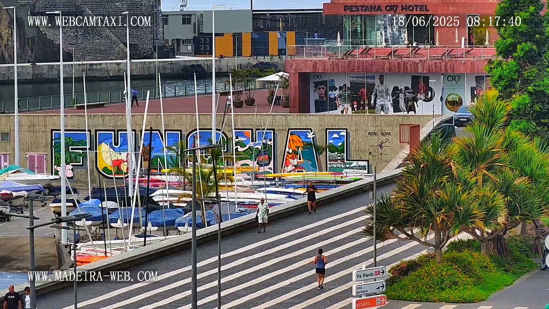 A harbor filled with boats is next to a building with a large mural that says "FUNCHAL," and people are walking on a paved area, under a sunny sky.