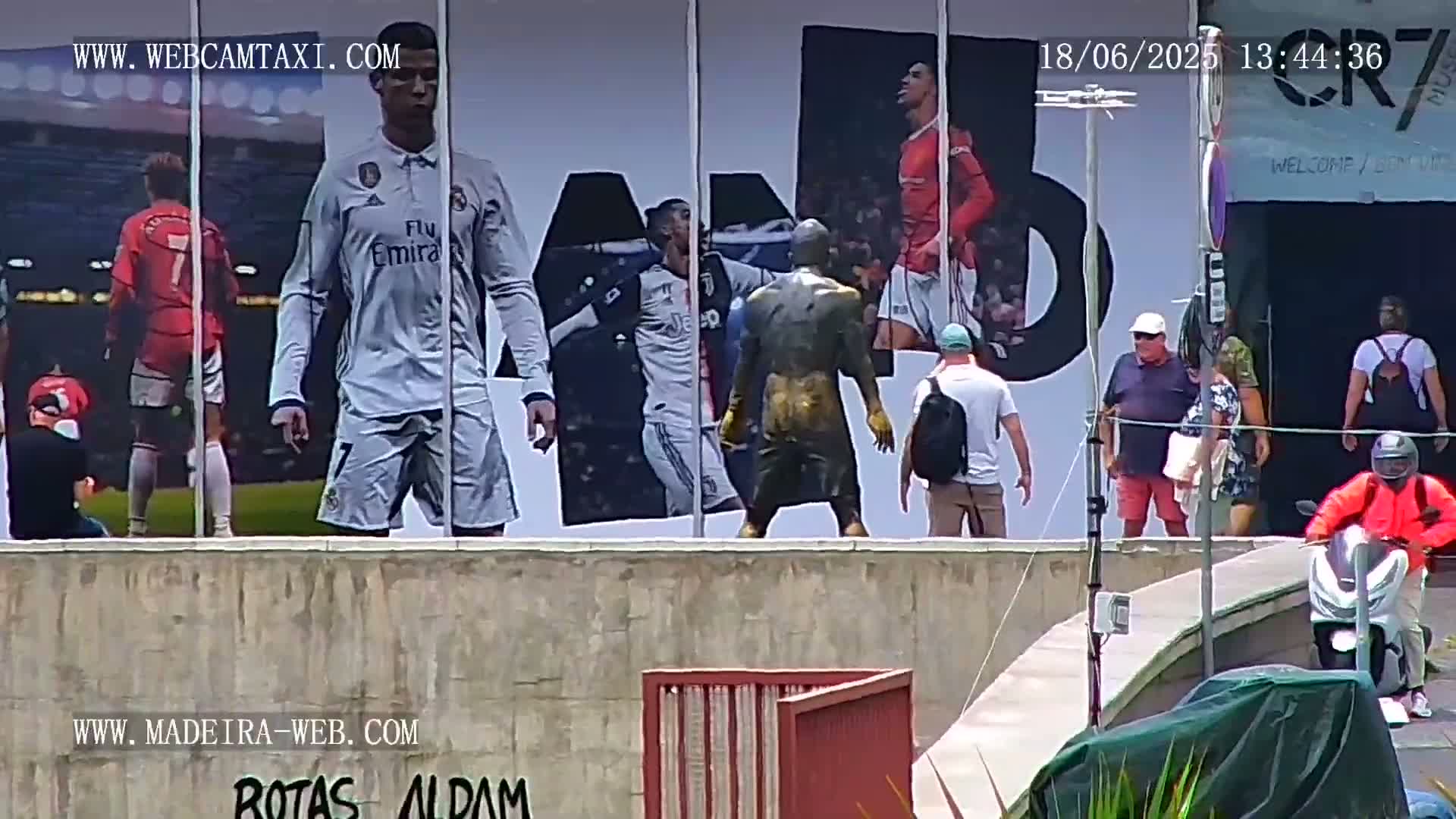 A large wall displays images of Cristiano Ronaldo in different soccer jerseys, alongside a bronze statue of a soccer player, while several people walk by on a sunny day.