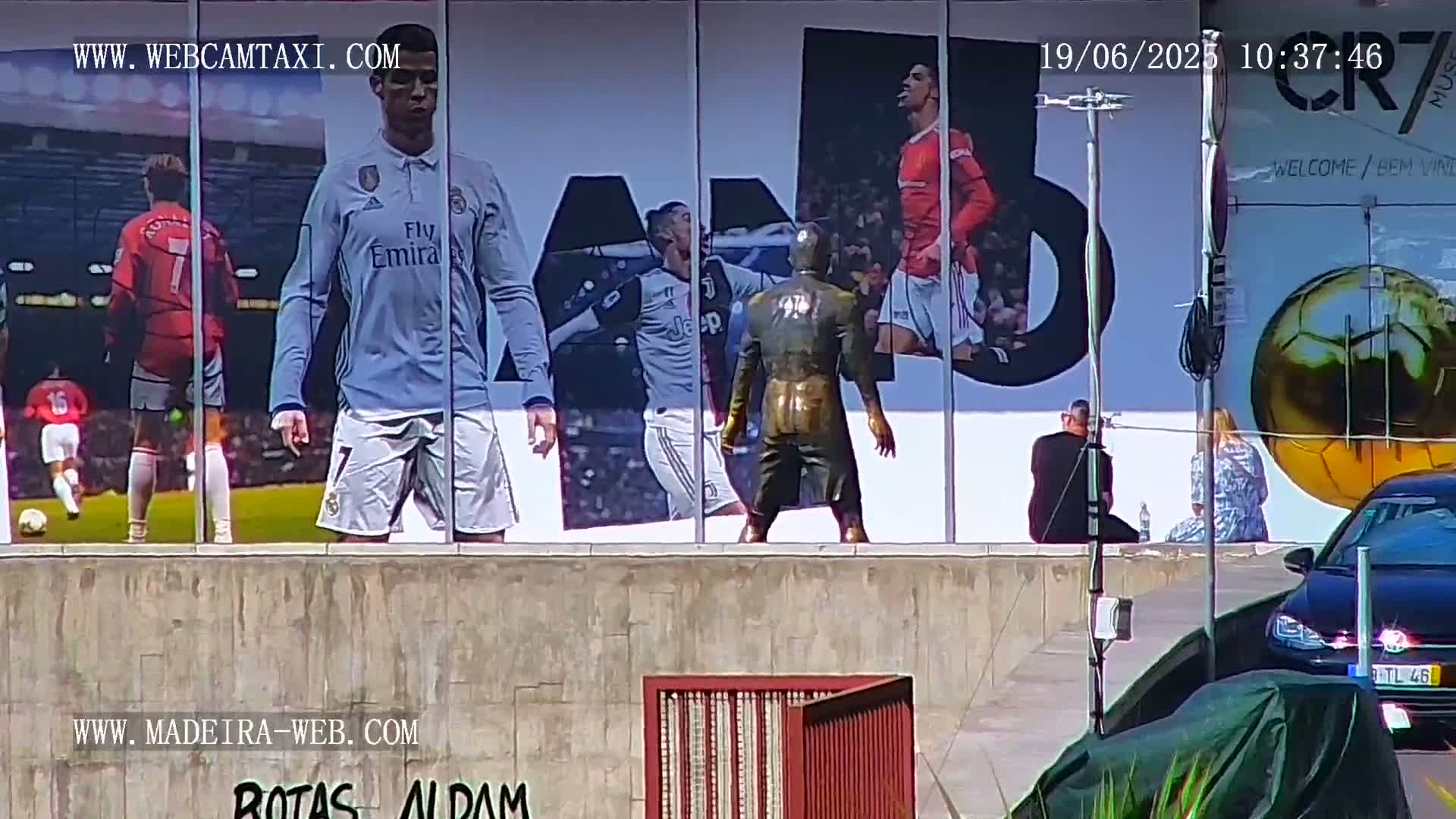 A large billboard depicting Cristiano Ronaldo in various soccer uniforms is visible on a sunny day, with a bronze statue of a soccer player in front of it and a few people sitting nearby.