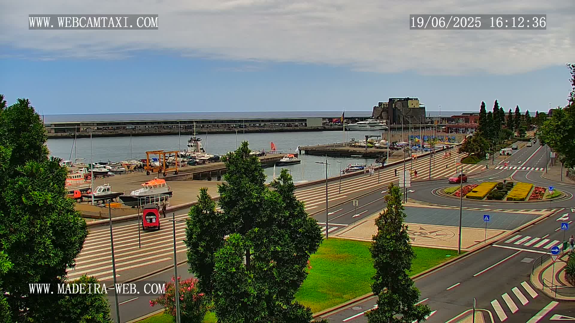 A harbor filled with boats is visible from a high vantage point on a partly cloudy day, with roads and green spaces leading away from the waterfront.