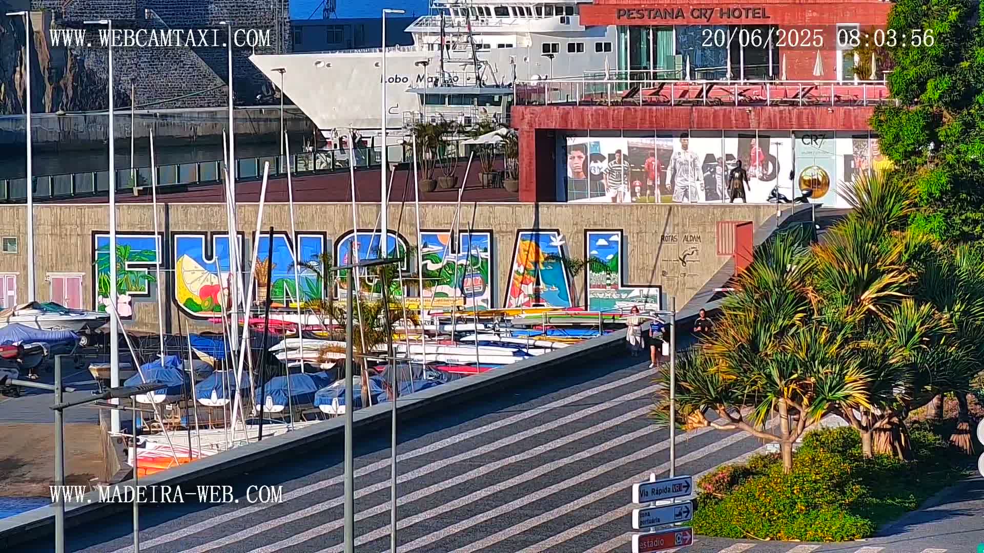 A harbor scene on a sunny day shows numerous small sailboats docked, a large mural depicting the word "Funchal," a cruise ship in the background, and pedestrians walking along a striped walkway.