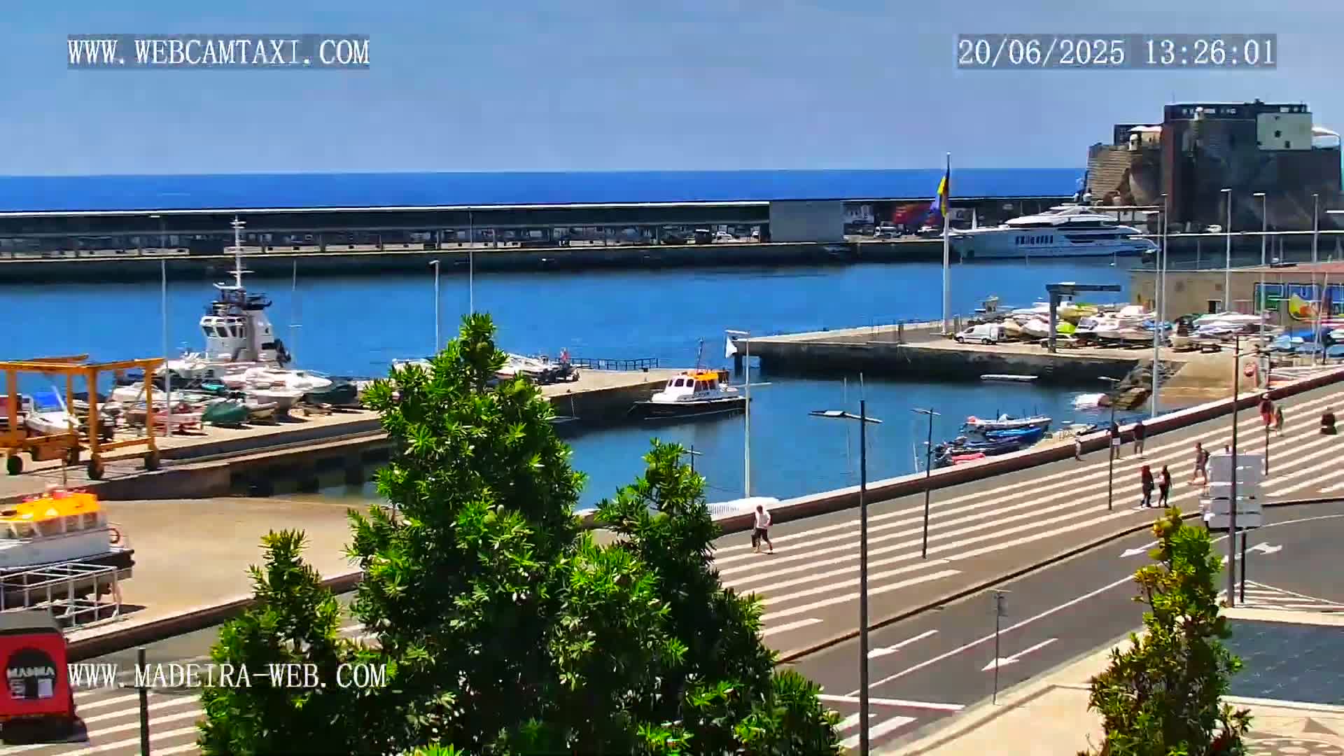 A sunny harbor scene shows numerous boats docked, several people walking along a paved waterfront, and a larger vessel in the water beyond the dock.