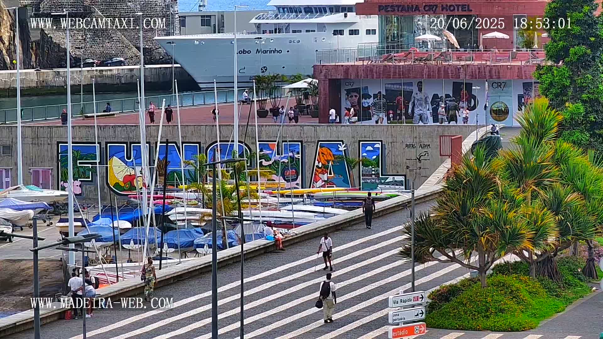 A harbor scene on a sunny day shows a large mural of the word "Funchal" on a wall next to a marina filled with boats, with pedestrians walking along a striped walkway and a large cruise ship in the background.