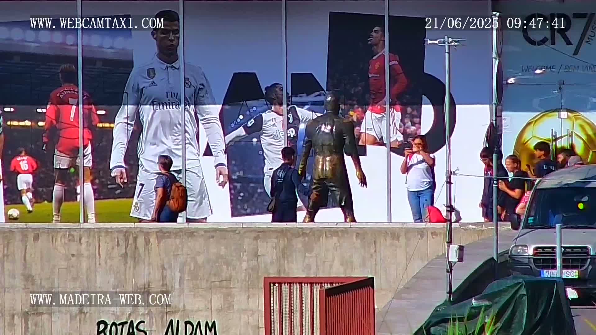 A large building displays a billboard featuring images of Cristiano Ronaldo in different soccer team uniforms, alongside several people standing in front of it on a sunny day.