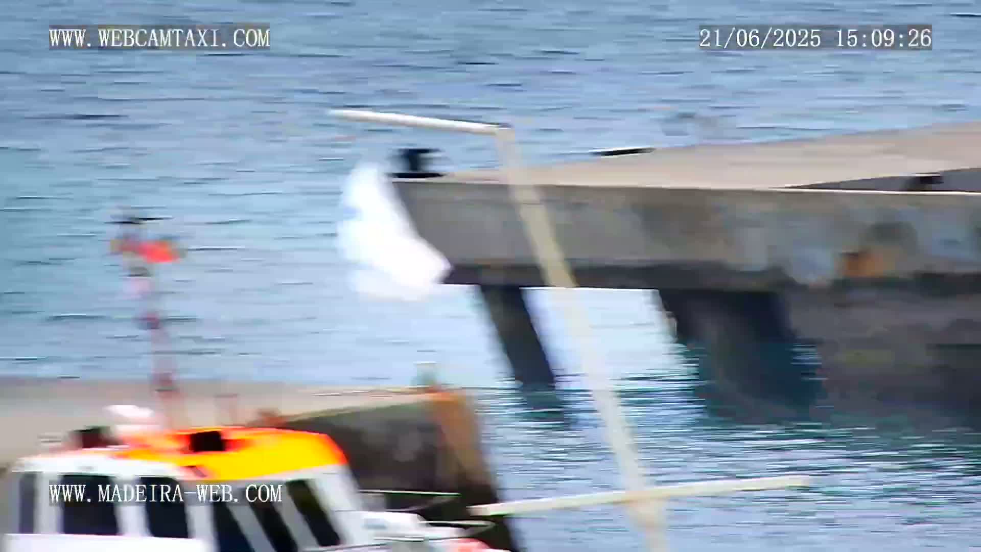 A partially visible orange and white boat is near a concrete dock in calm, blue water, with a white cloth waving in the breeze.