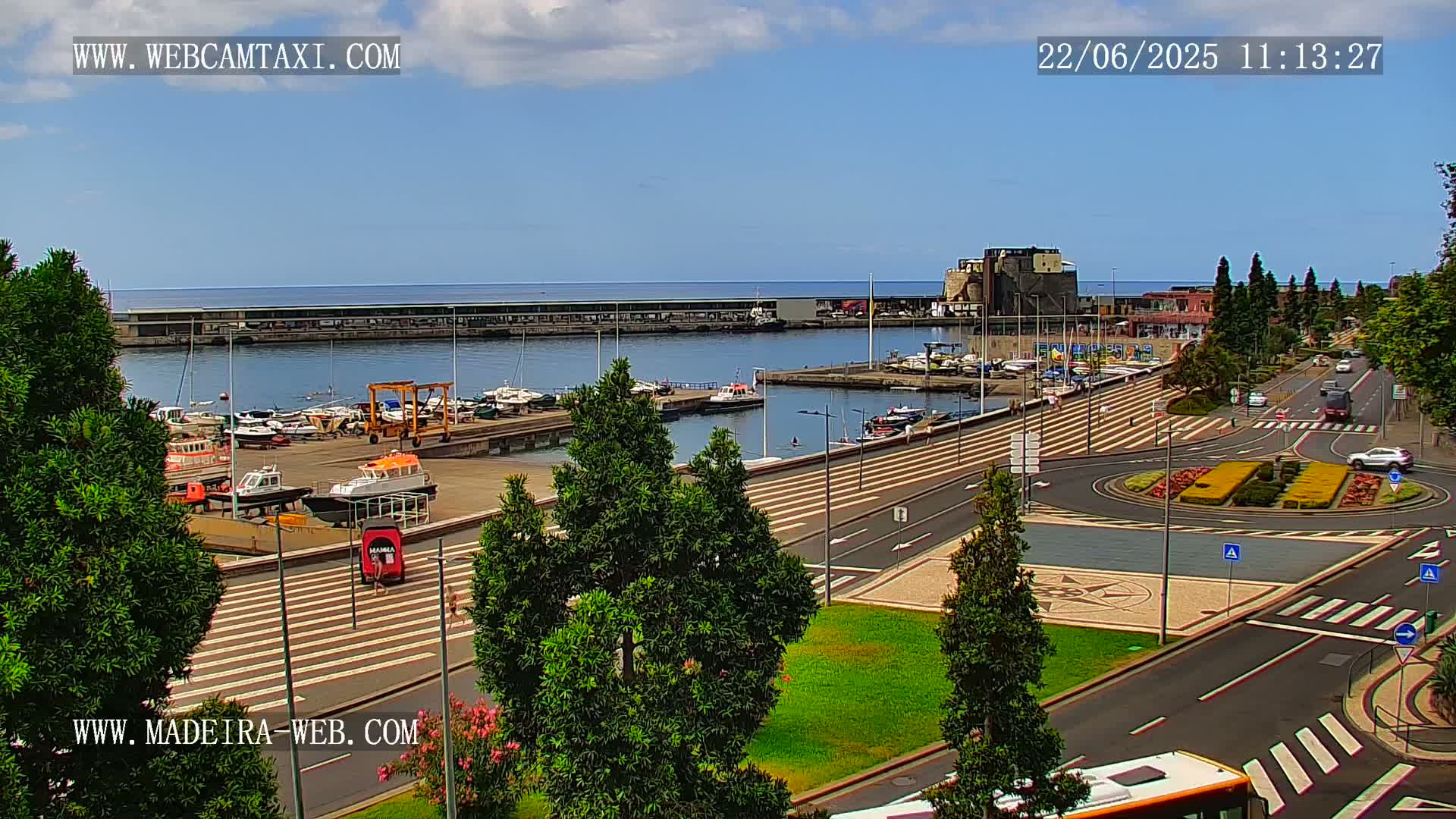 A harbor filled with boats is visible from a high vantage point on a sunny day, overlooking a paved plaza with a roundabout and a road with cars.