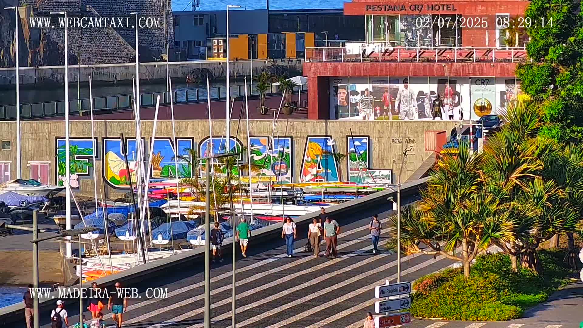 A harbor scene with numerous sailboats and motorboats docked along a wall depicting a colorful mural of the word "Funchal," under sunny skies, and several people walking on a striped pedestrian walkway.