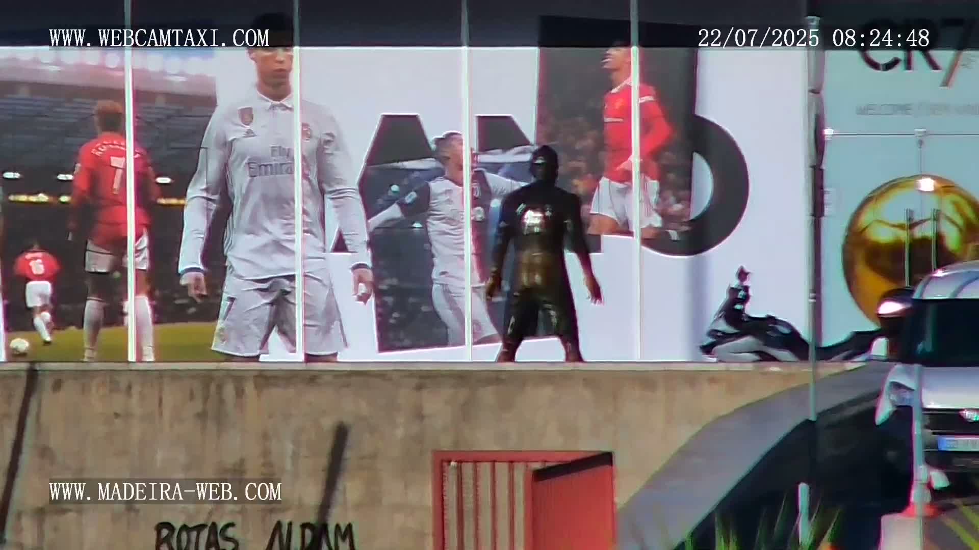 A large building displays images of soccer players, while a dark figure stands in front of it on a sunny day.