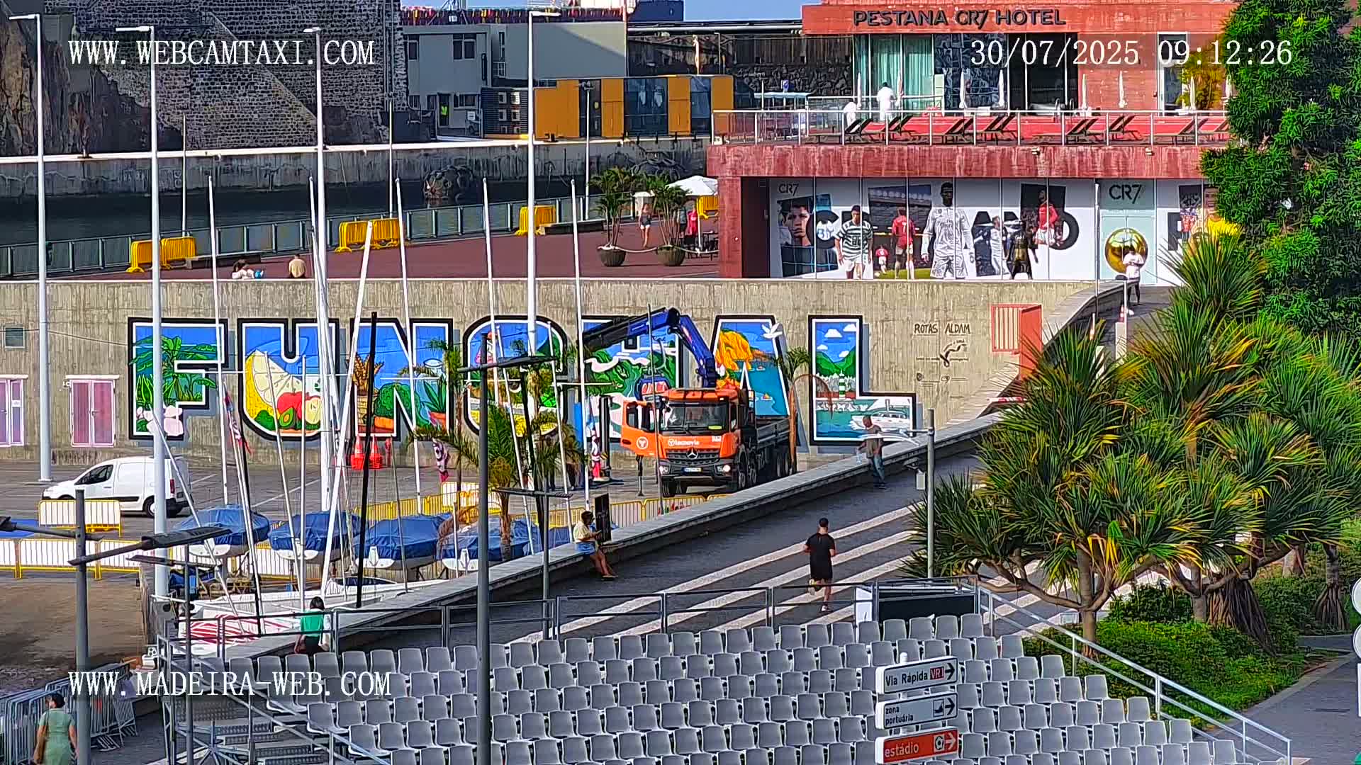 A sunny day shows a harbor scene with a large mural, a small orange truck with a crane, several people walking around, and empty stadium seating in the foreground.
