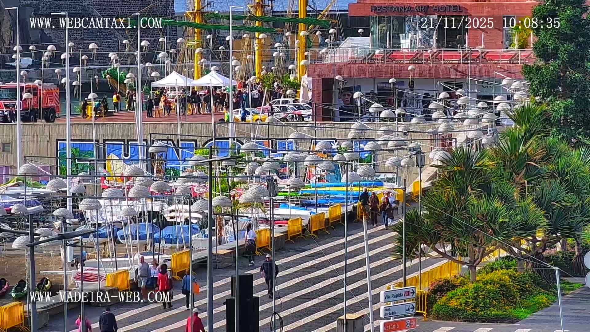 Under bright sunshine, a bustling harbor area is adorned with numerous jellyfish-shaped light fixtures hanging overhead, showcasing people walking, a red emergency vehicle, rally cars, a tall ship, and a prominent hotel building in the background.