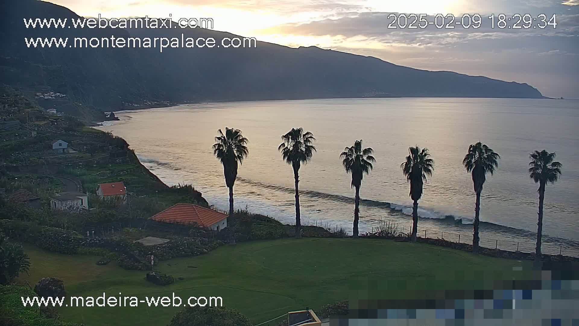 A coastal scene at sunset shows a calm ocean with small waves lapping the shore, several palm trees silhouetted against the water, and a few houses nestled on a hillside overlooking the sea.