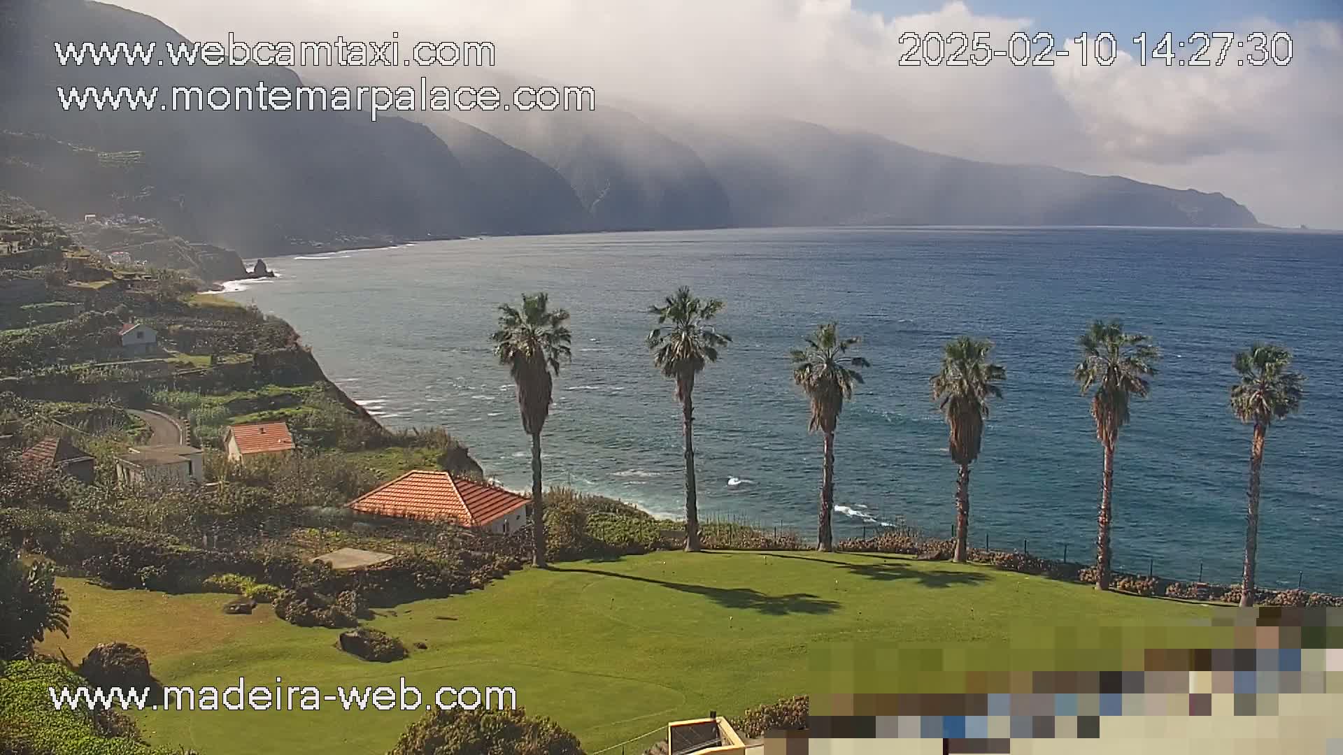 A partly cloudy day shows a coastal scene with a calm ocean, palm trees lining a grassy area near buildings and cliffs, and mountains in the distance.