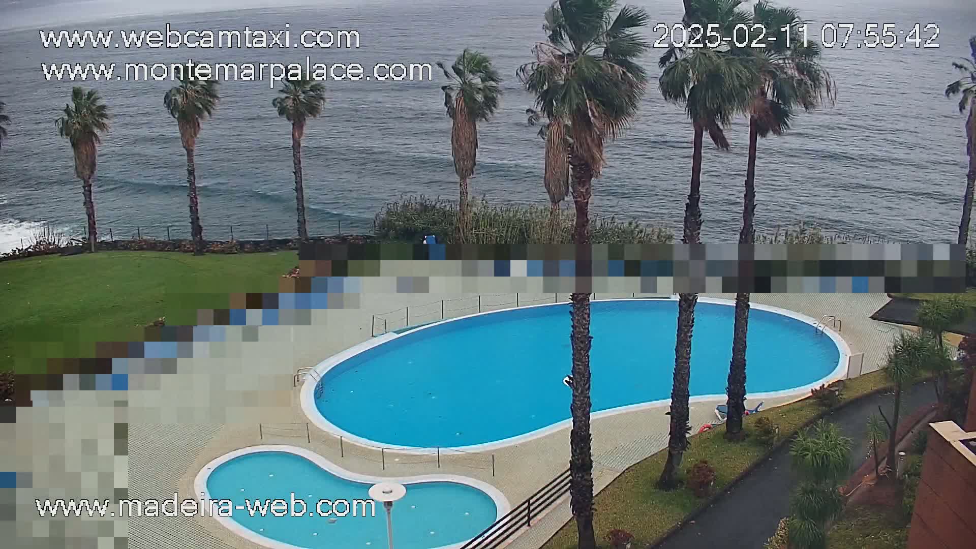 An outdoor kidney-shaped swimming pool and a smaller adjacent pool are situated on a grassy area overlooking a wavy ocean under an overcast sky.