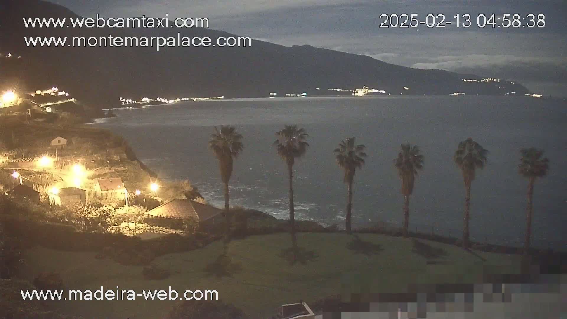 A nighttime coastal scene shows a dark ocean, a hill with houses and palm trees, and distant mountains under a cloudy sky.
