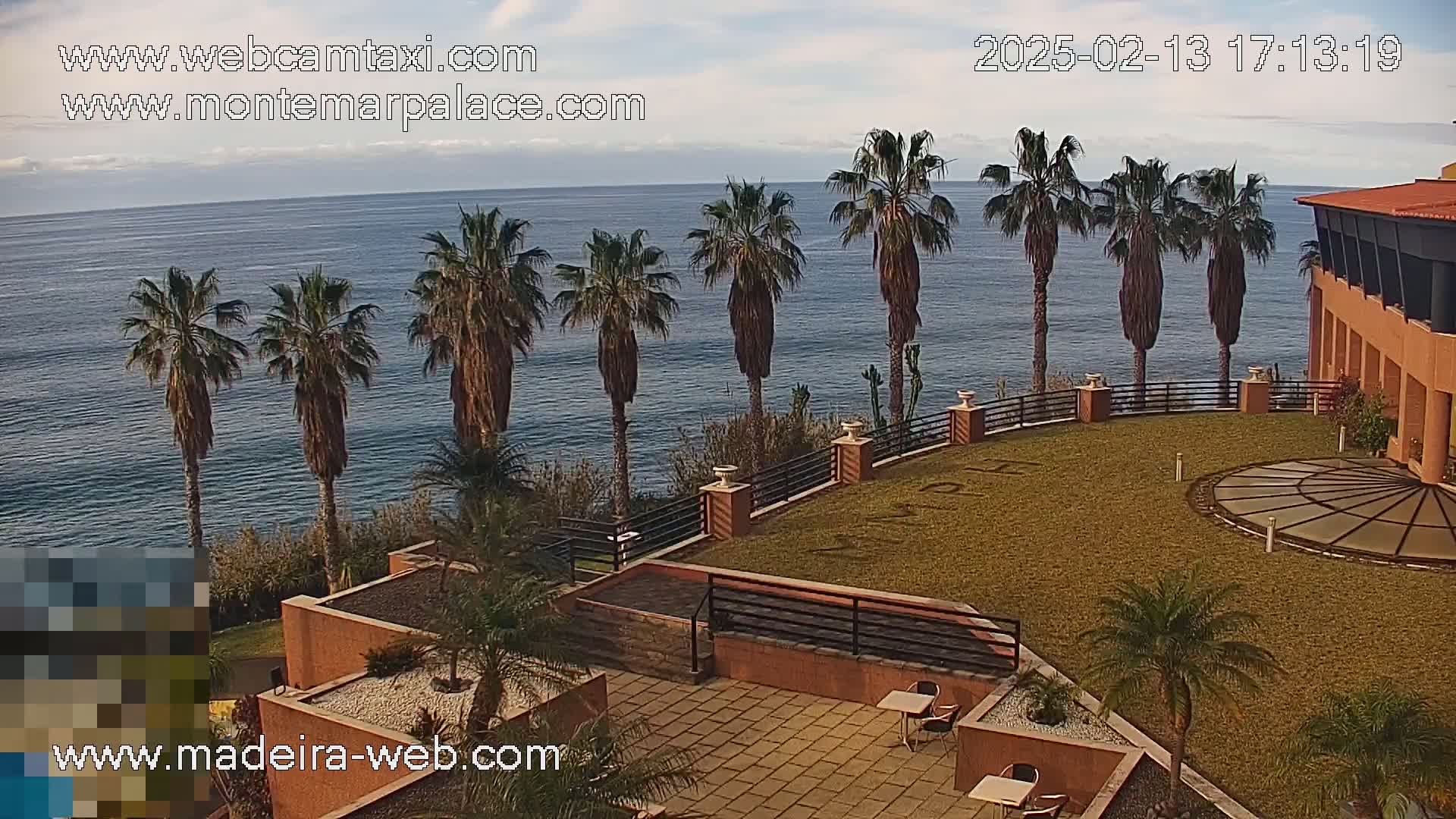 A partly cloudy day overlooks a resort's patio and lawn with palm trees, a circular paved area, and a view of the ocean.