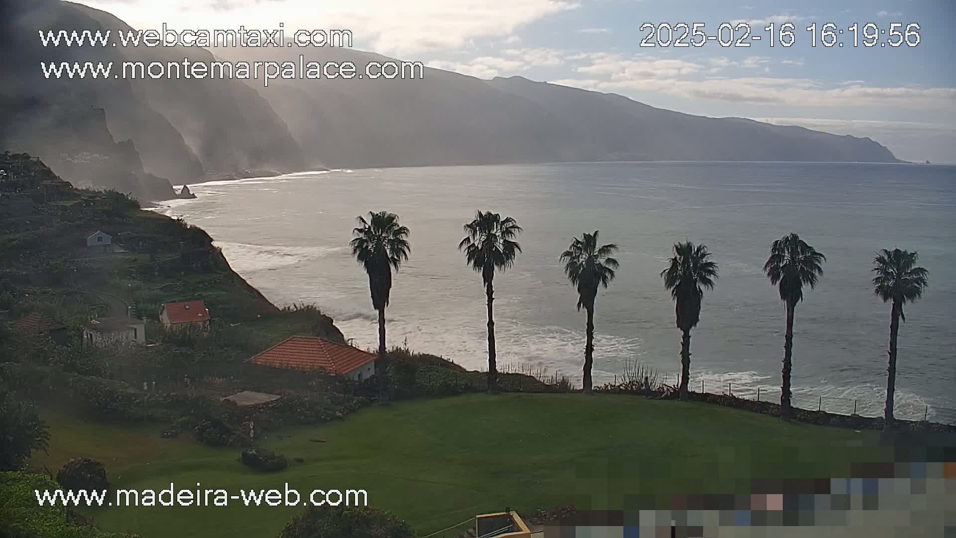 A coastal scene under partly cloudy skies shows a rocky shoreline with houses, palm trees, and a calm ocean with small waves.