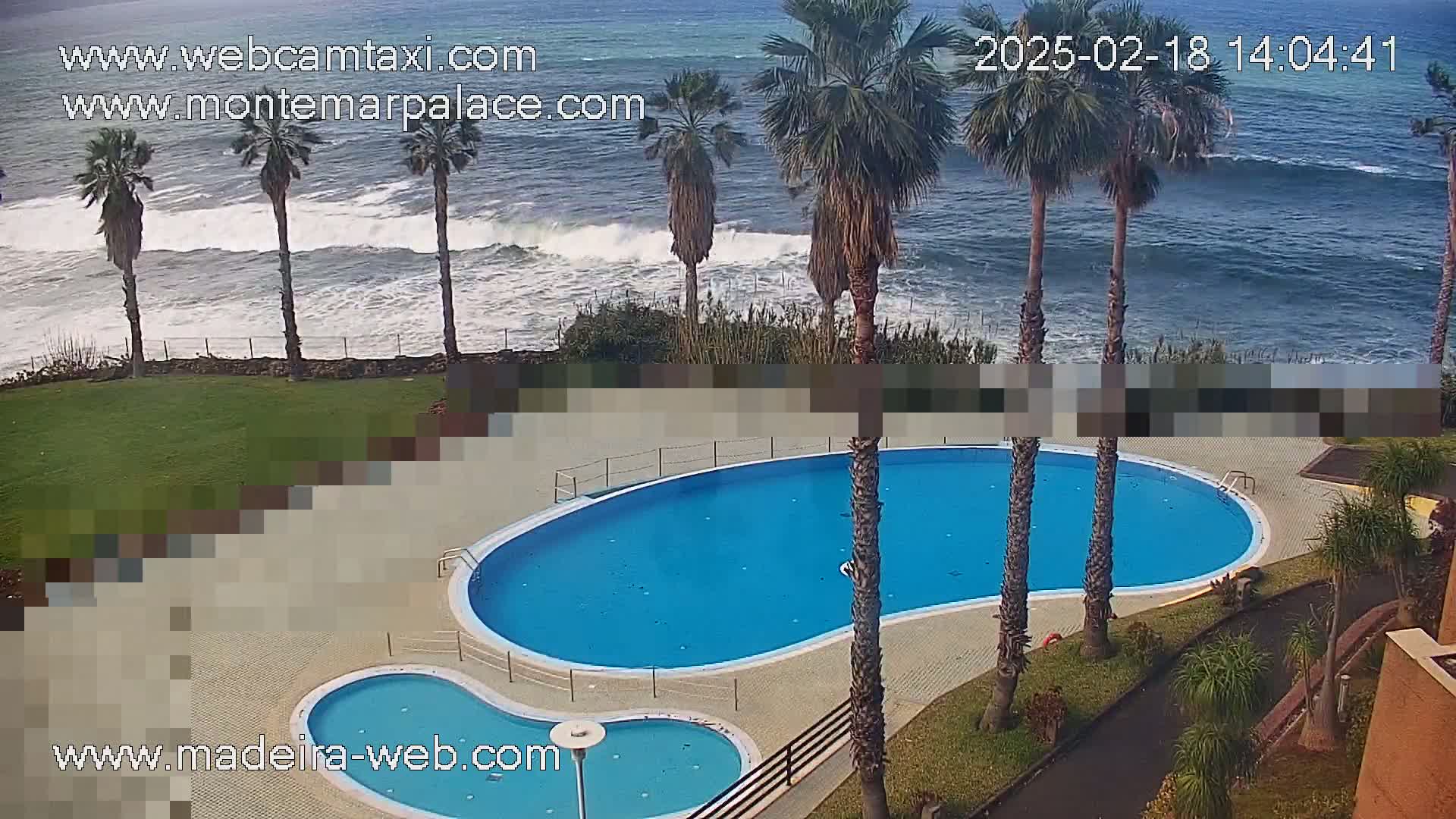 An ocean-side kidney-shaped swimming pool with a smaller children's pool next to it, surrounded by palm trees and overlooking a wavy ocean under a somewhat cloudy sky.