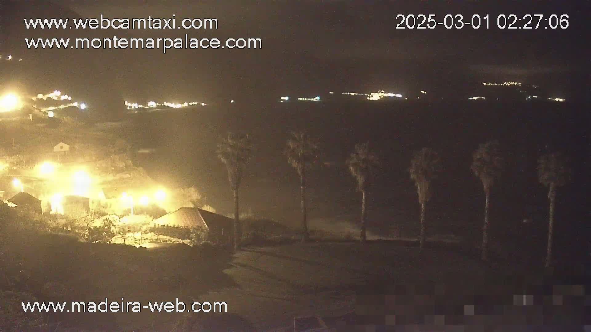 A nighttime coastal scene shows a village with illuminated buildings and palm trees, with waves crashing against the shore under a dark sky.