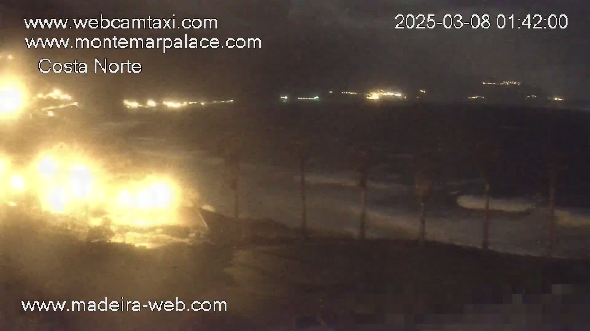 A nighttime coastal scene shows bright headlights of vehicles on a road, with waves crashing on the shore under a dark, cloudy sky.