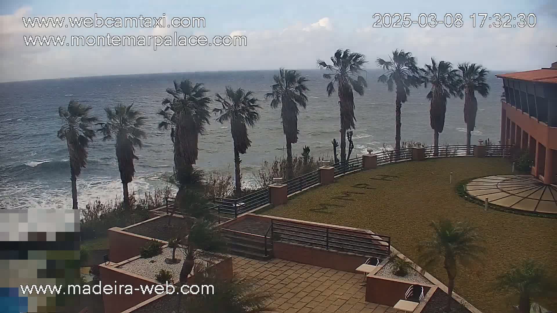 A partly cloudy day shows a rocky ocean shore with palm trees, a building with a courtyard, and a circular paved area.