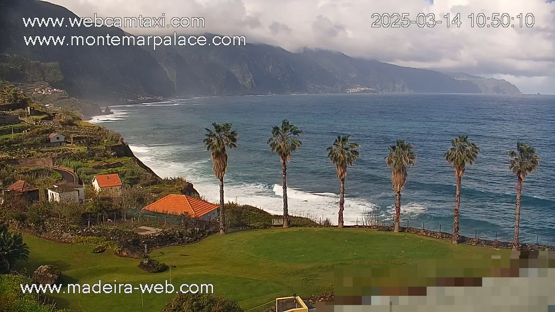 A partly cloudy day shows a coastal scene with a rocky, green hillside dotted with houses, several palm trees on a grassy area overlooking a dark blue ocean with small waves breaking on the shore, and mountains in the background.