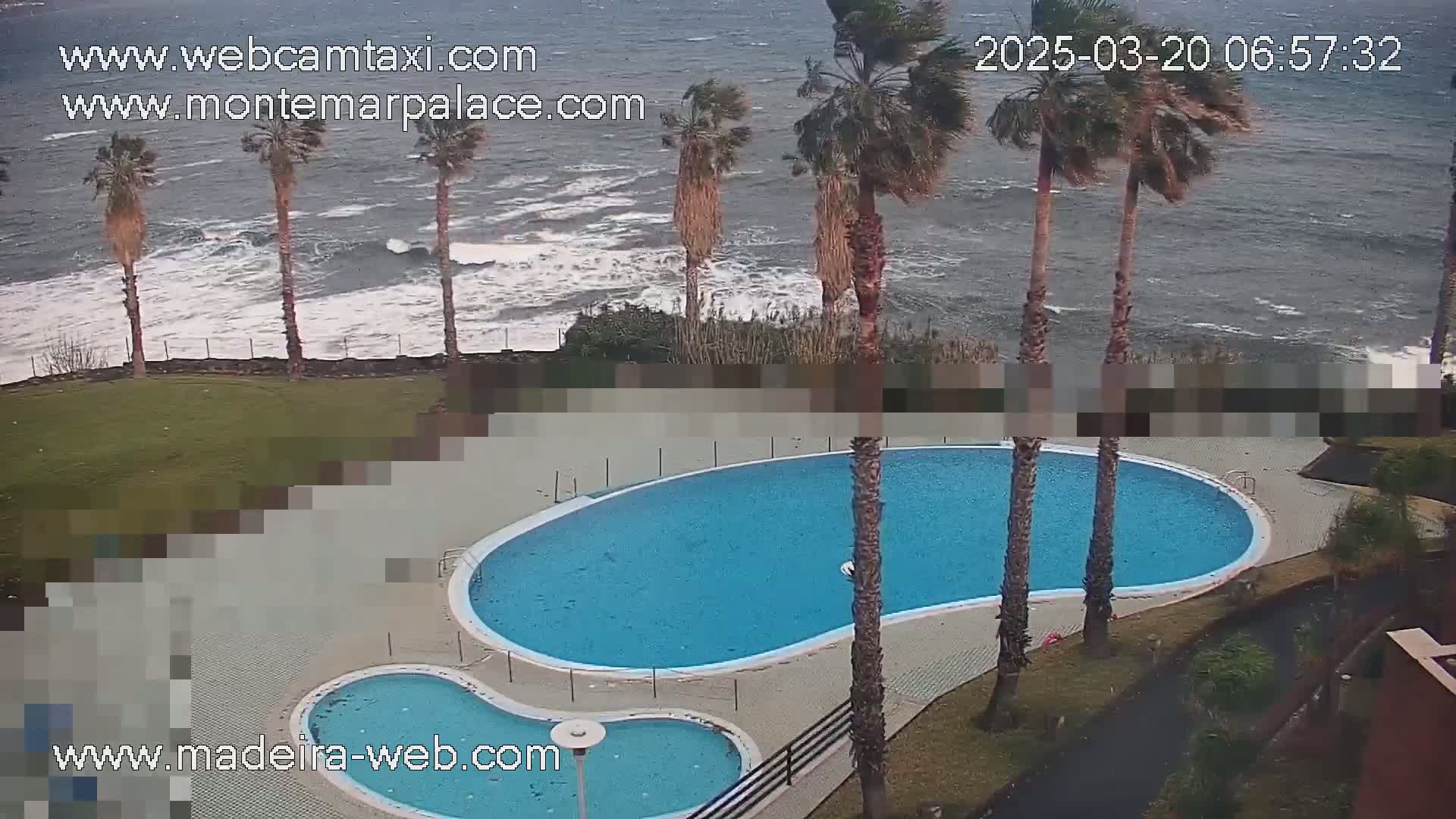 An outdoor swimming pool complex next to a wavy ocean with palm trees under a cloudy sky.