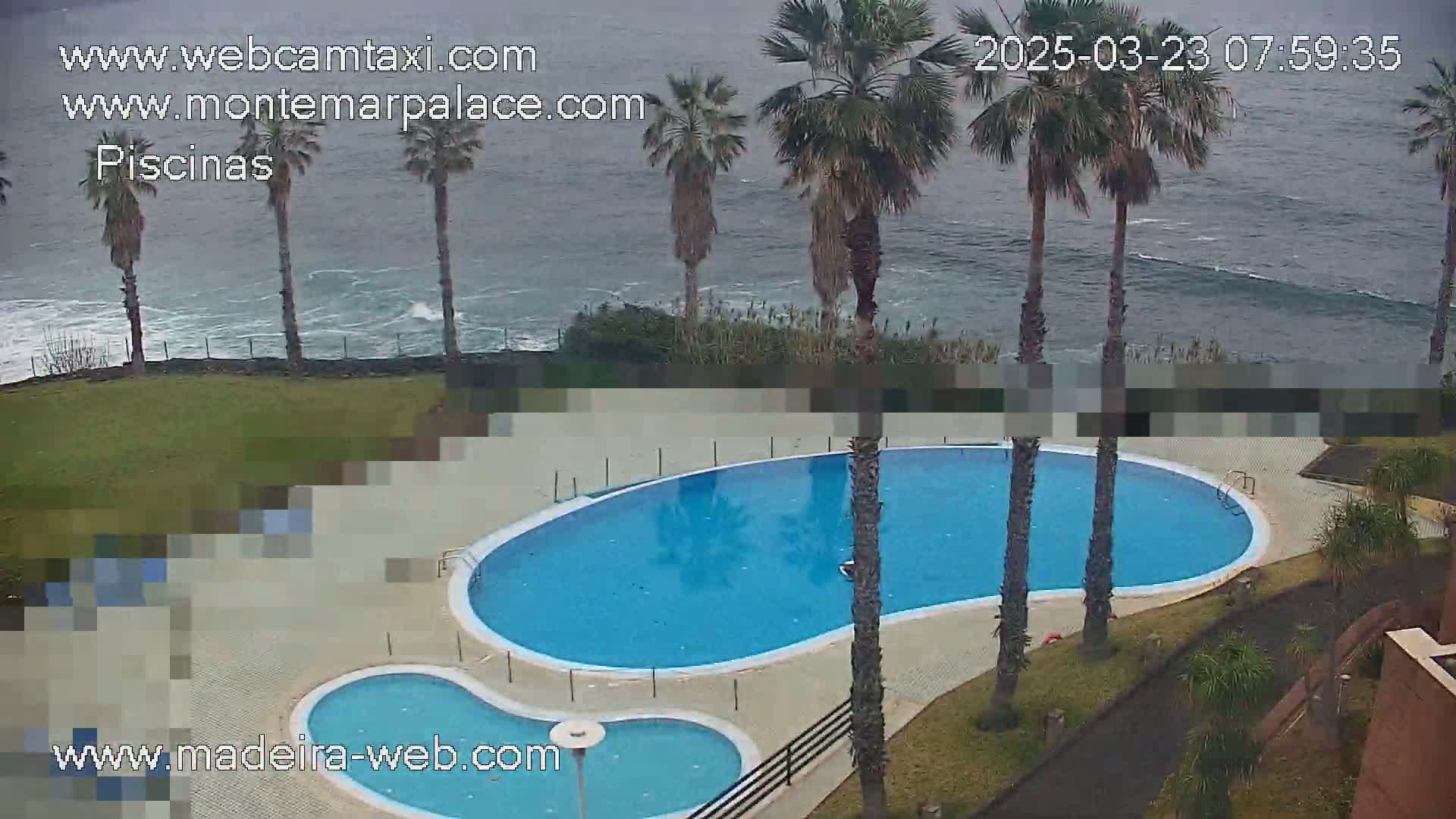 An outdoor kidney-shaped swimming pool and a smaller adjacent pool are situated next to a grassy area overlooking a wavy ocean under an overcast sky.
