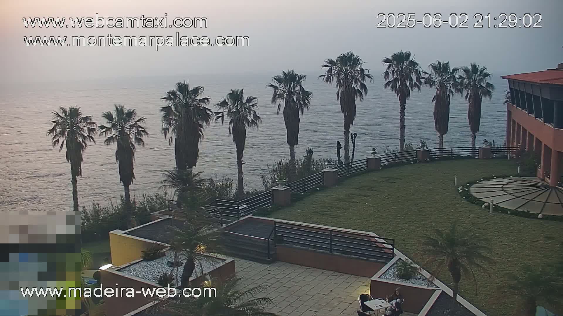 A calm ocean is visible behind a resort with palm trees and manicured lawns, under a hazy evening sky.