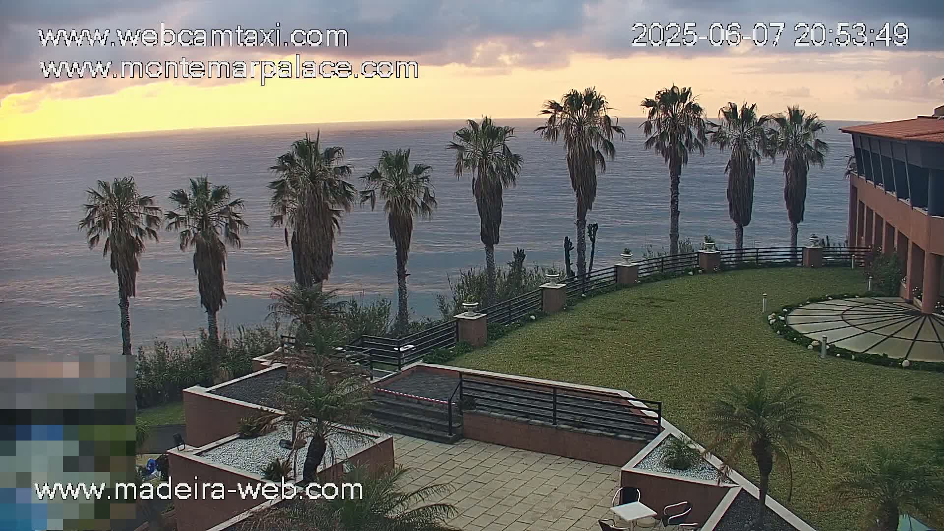 A coastal resort with palm trees overlooks a calm ocean at sunset under a partly cloudy sky.
