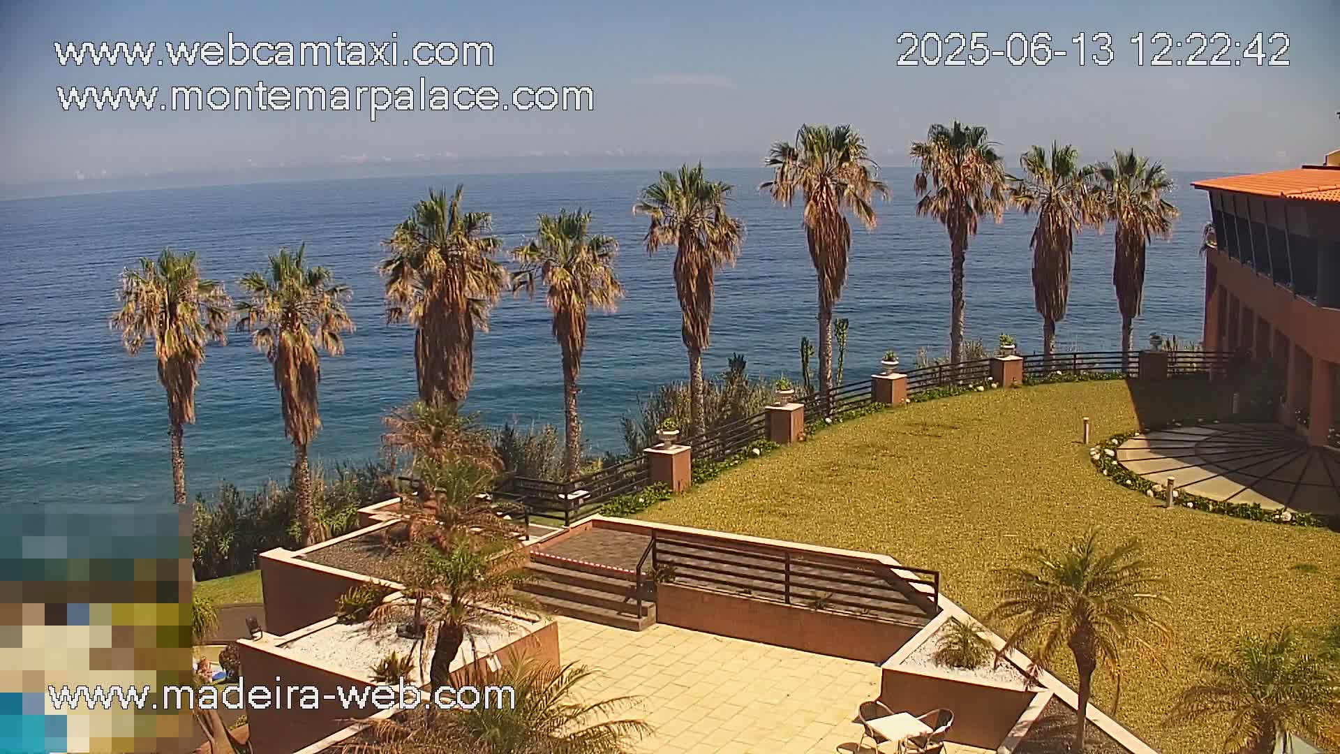 A sunny ocean view from a cliffside property features palm trees, a manicured lawn, and a building with terracotta-colored exterior.
