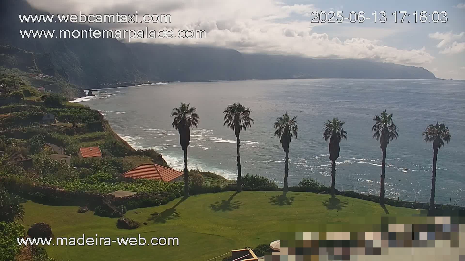 A partly cloudy day shows a tranquil ocean scene with a rocky coastline, terraced hillsides with small buildings, and several palm trees on a grassy lawn overlooking the water.