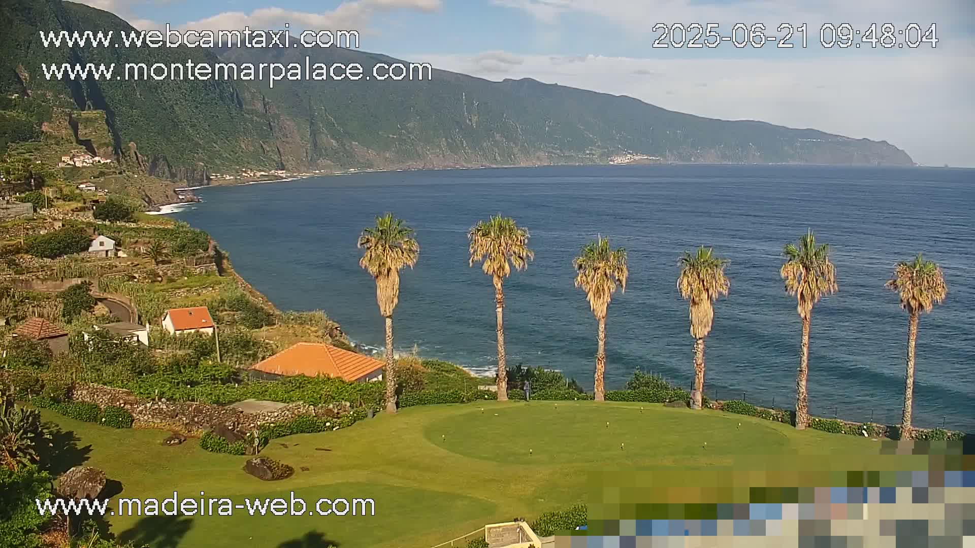 A putting green overlooks a calm ocean next to a mountainous coastline, under a mostly clear sky.