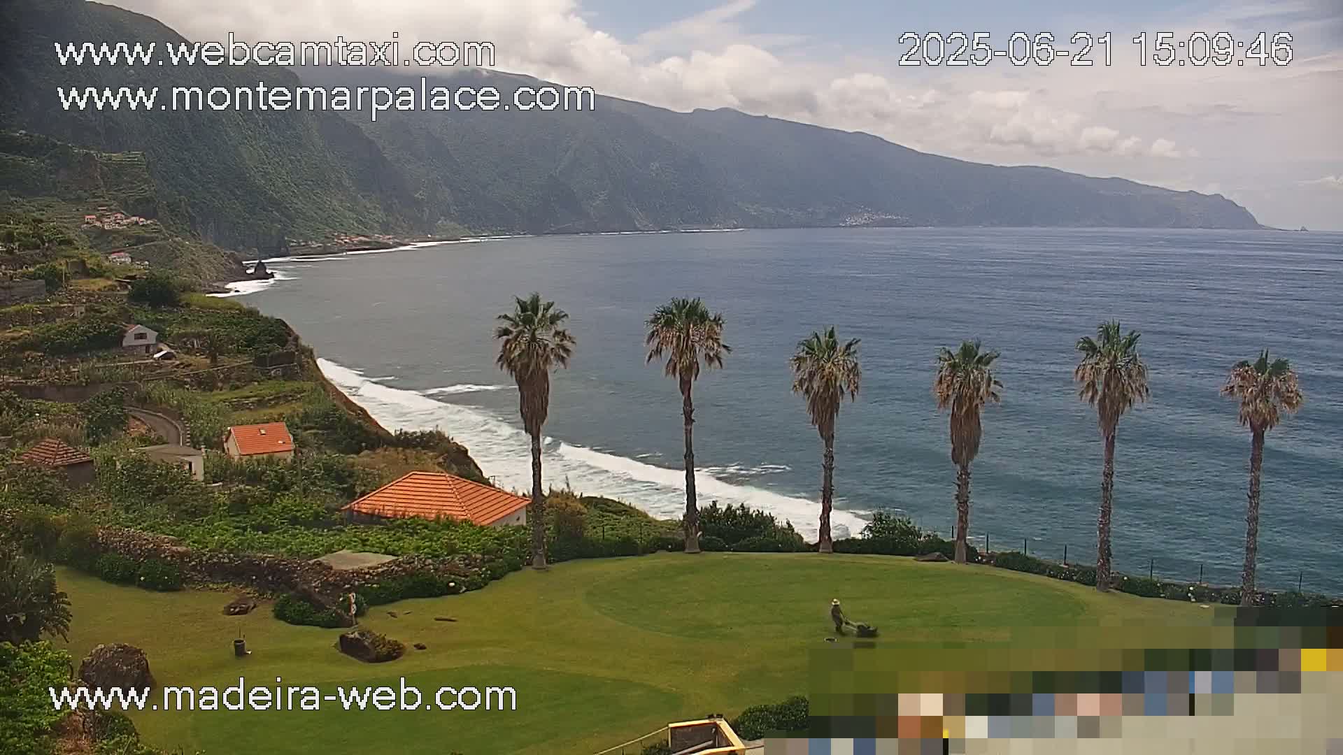 A person mows a lawn overlooking a calm ocean with waves breaking on a rocky shore, backed by a green hillside under a partly cloudy sky.