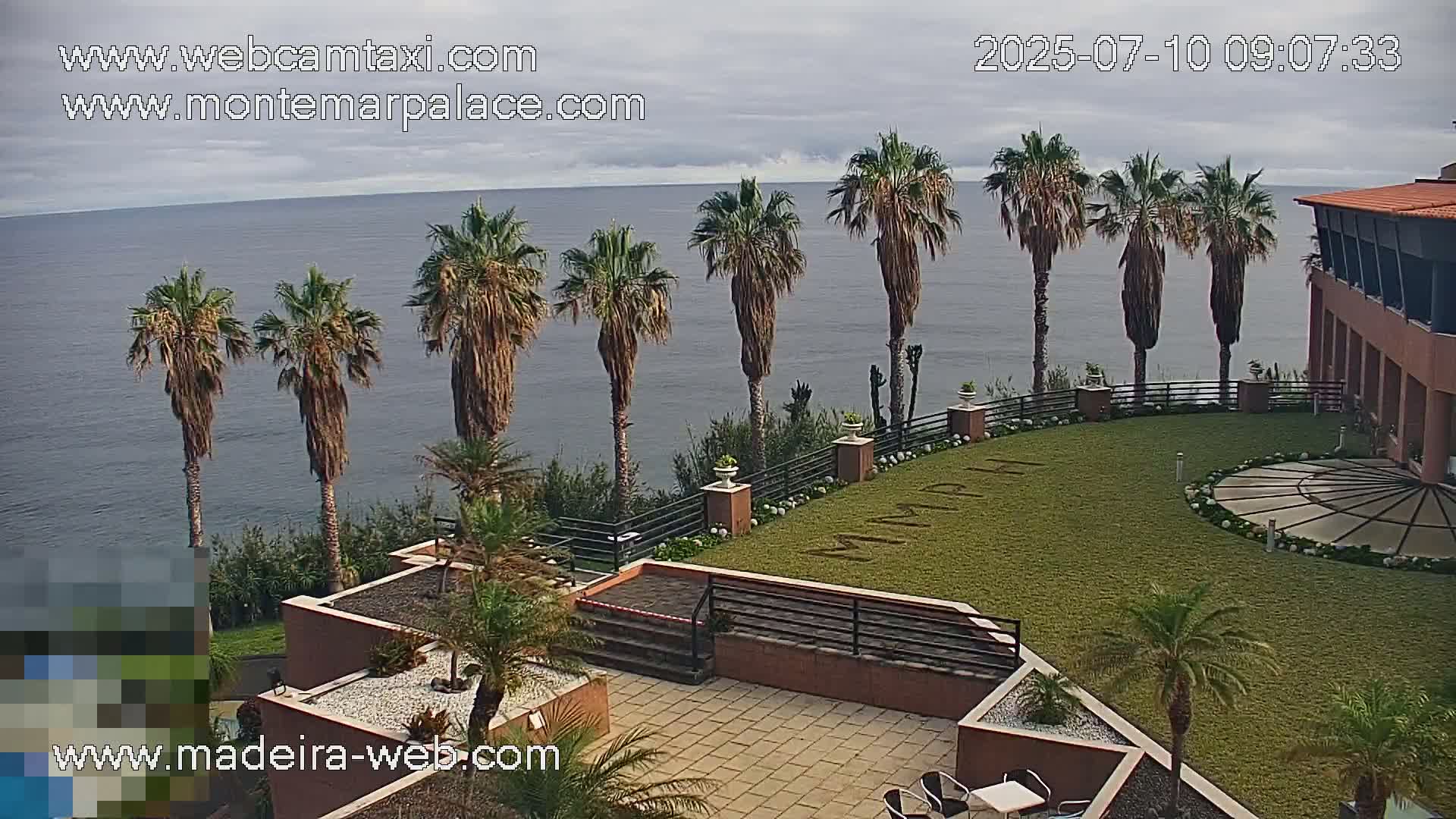 Overlooking a calm ocean, palm trees line a grassy courtyard with a circular patio area adjacent to a building under a cloudy sky.