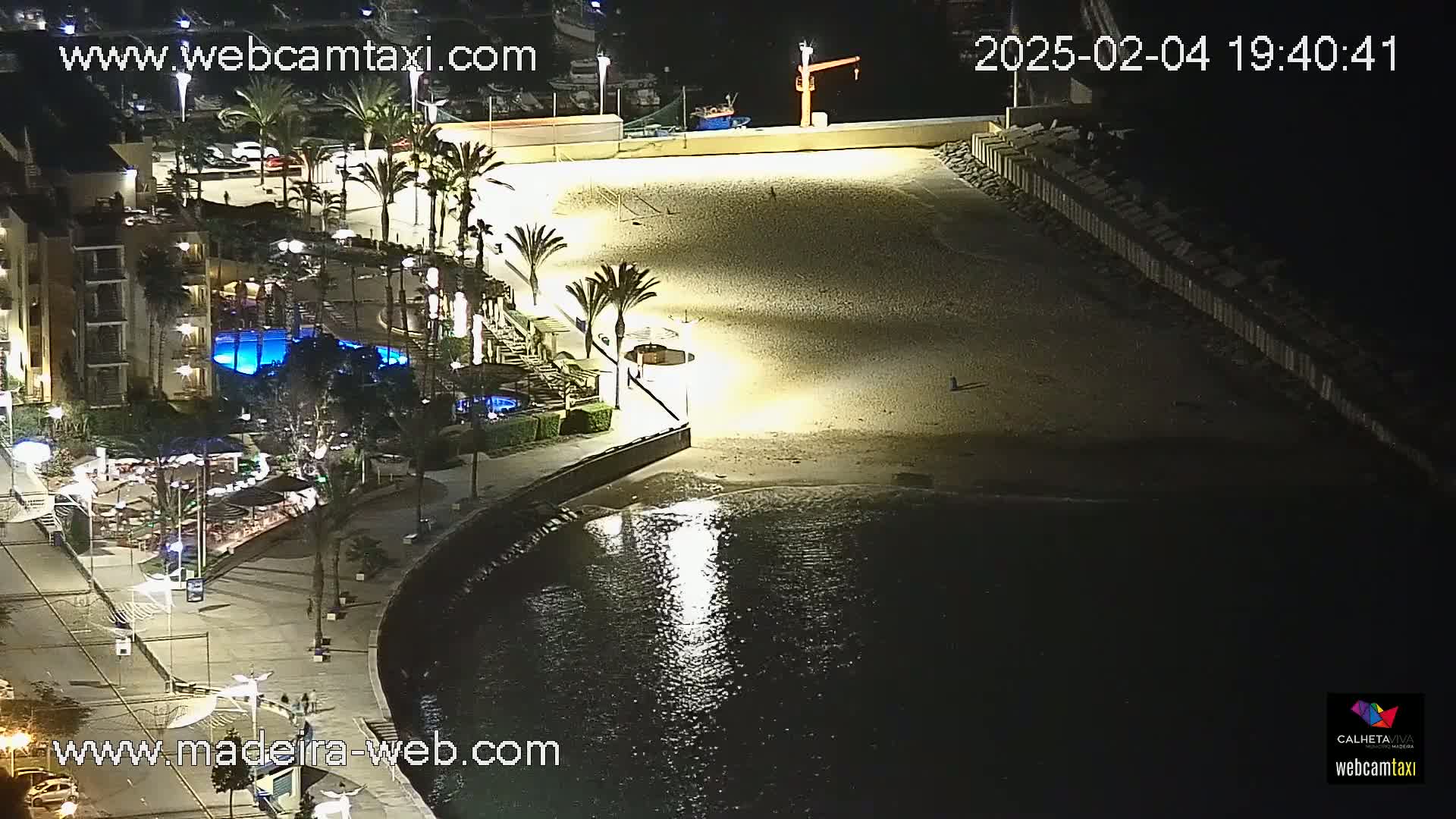 An illuminated beach at night, next to a building with palm trees and a pool, with calm dark water in front.