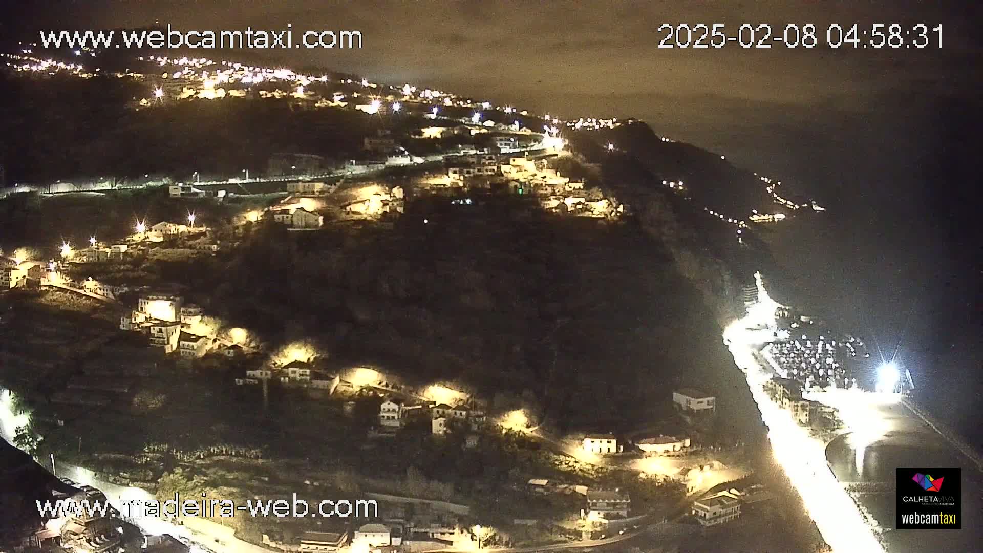 A nighttime, aerial view of a coastal town built on a hillside, with lights illuminating buildings and roads.