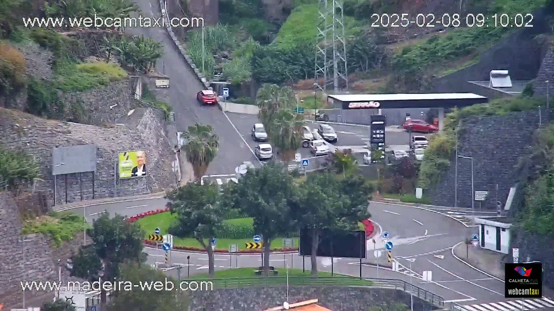 An aerial view shows a roundabout and a gas station surrounded by greenery and stone walls on an overcast day.