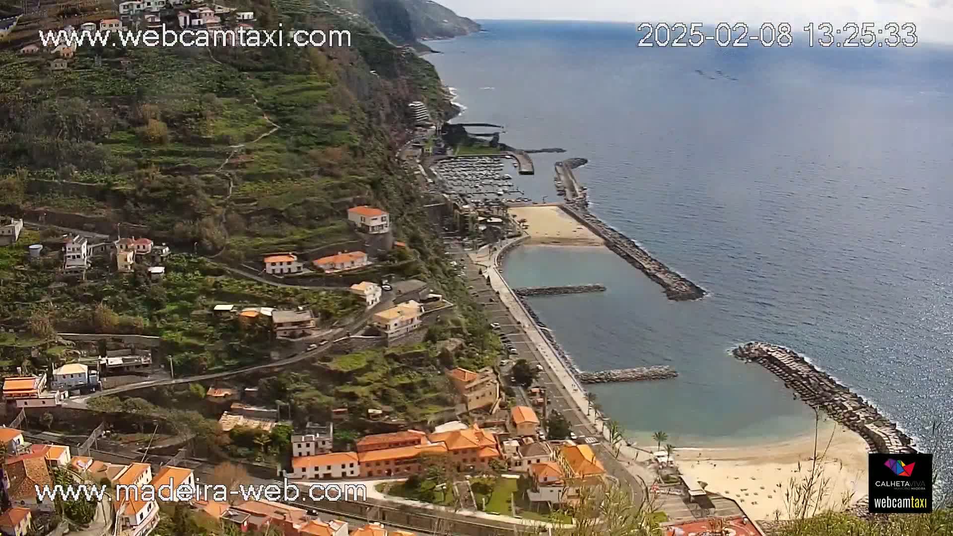 A coastal town nestled on a steep hillside overlooks a calm ocean with a small, sandy beach and a marina; the day is sunny.