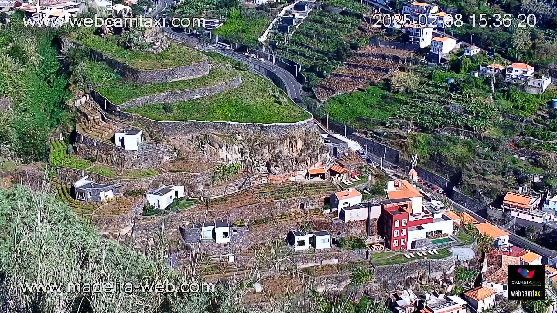 A hillside village with terraced gardens and houses overlooks a winding road on a sunny day.