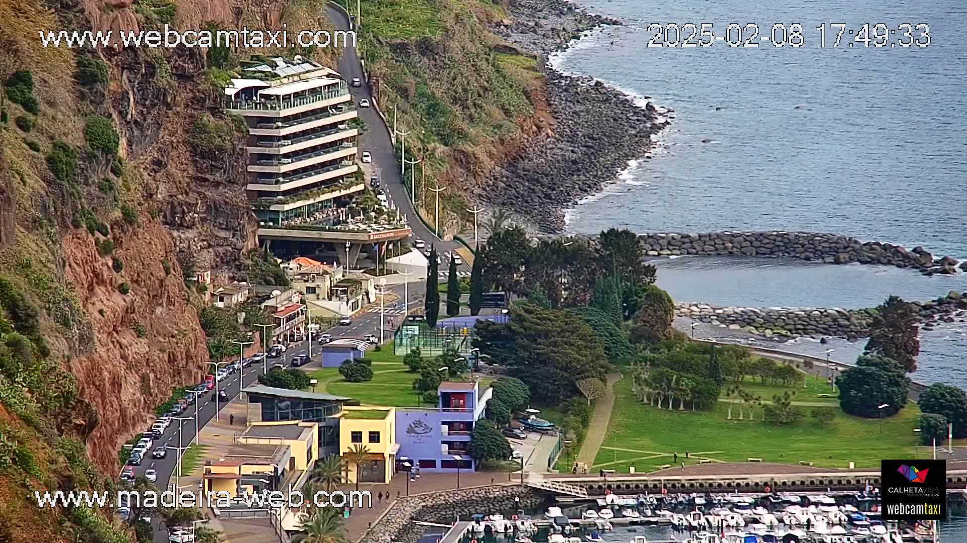 A coastal scene shows a multi-story building perched on a cliff overlooking a small harbor filled with boats, a park, and a calm ocean under a partly cloudy sky.