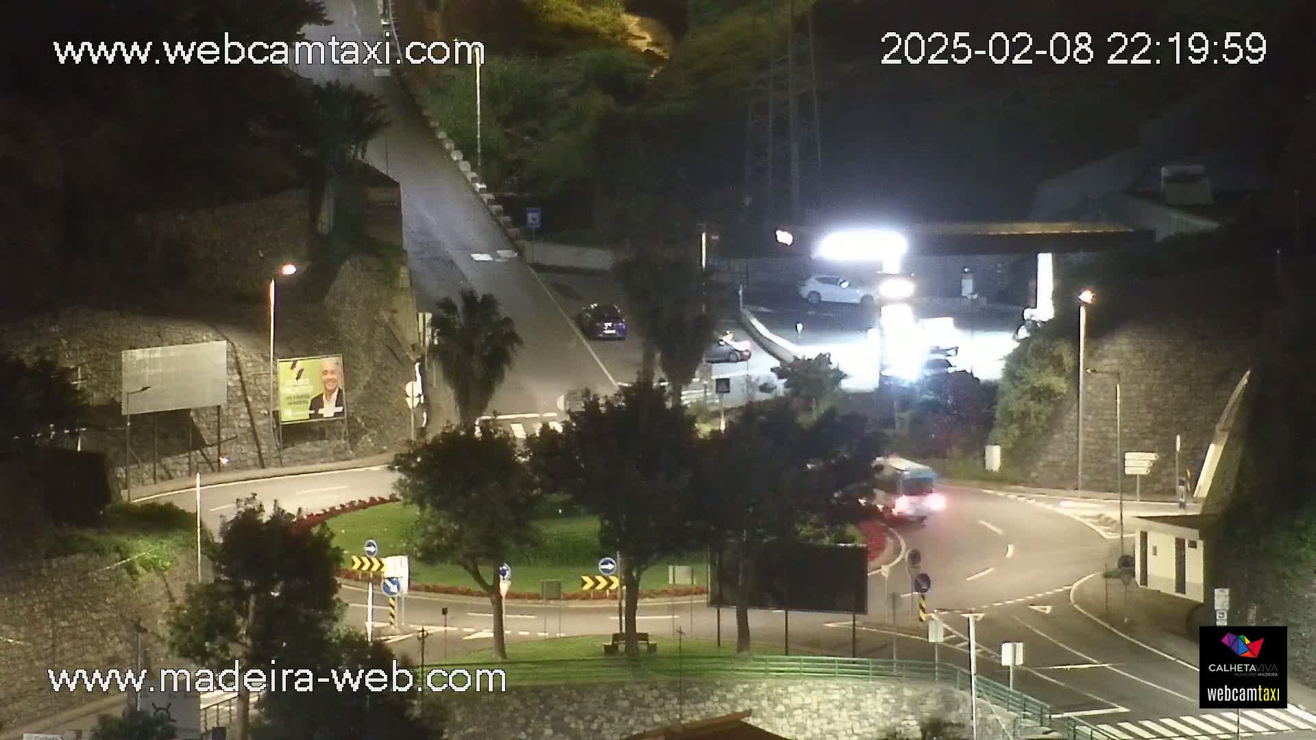 A nighttime view of a roundabout with several cars and a bus, situated near a gas station and surrounded by trees and stone walls under dark conditions.
