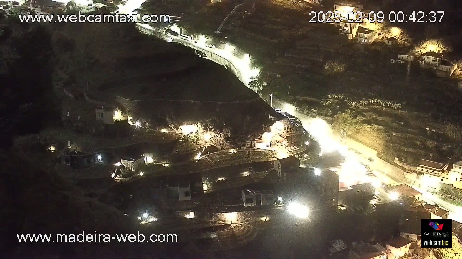 An aerial nighttime view of a coastal town shows a hillside with buildings illuminated by lights, a road winding through the area, and a cliff face partially visible.