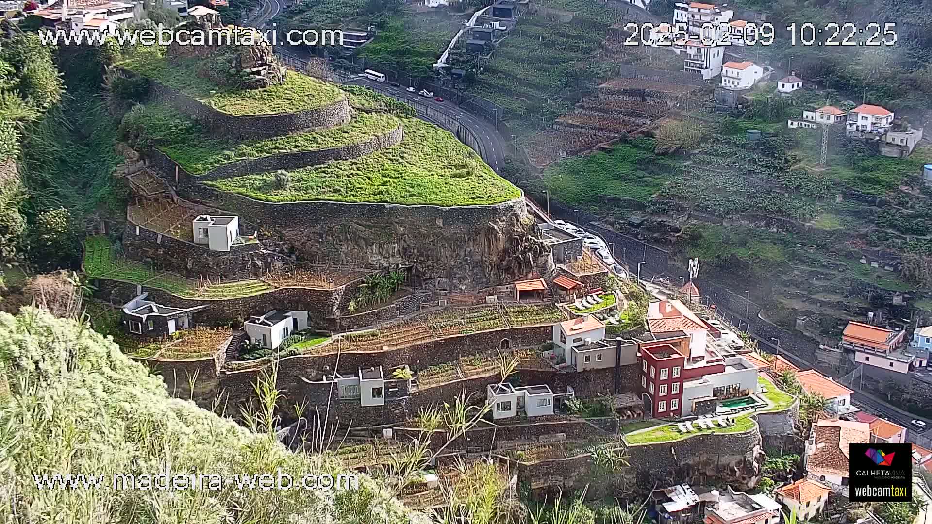 Terraced farmland and buildings cling to a hillside overlooking a road and other buildings, under a sunny sky.