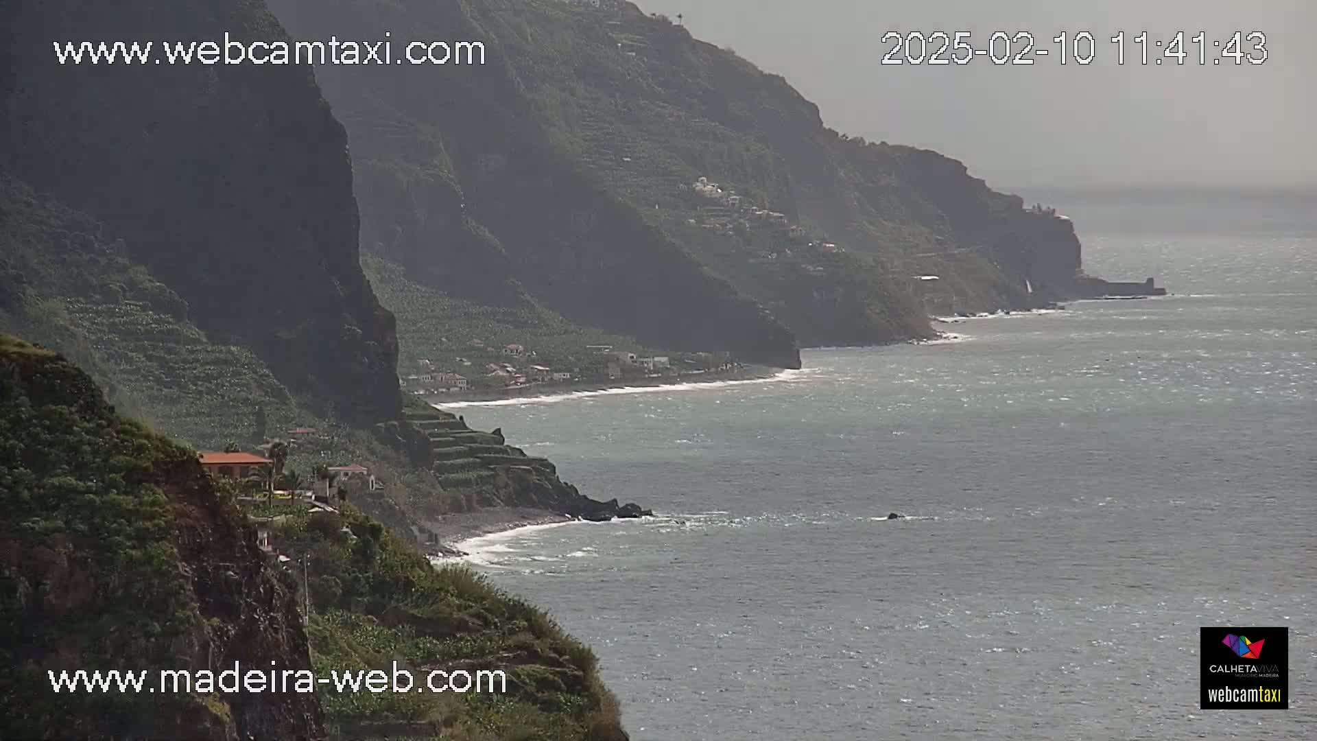 A hazy coastal scene shows a rocky, green cliffside with terraced farming, leading down to a pebble beach and a calm, gray ocean.